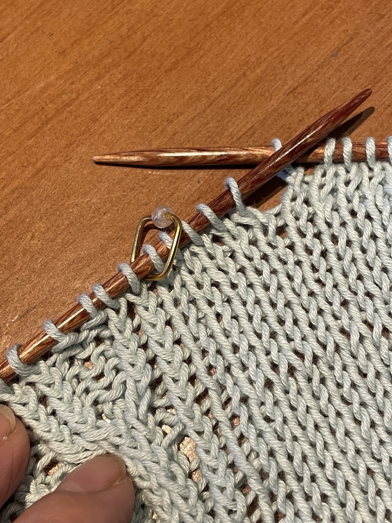 A knitting work in progress on the needles with a square-shaped handmade stitch marker sitting in between stitches