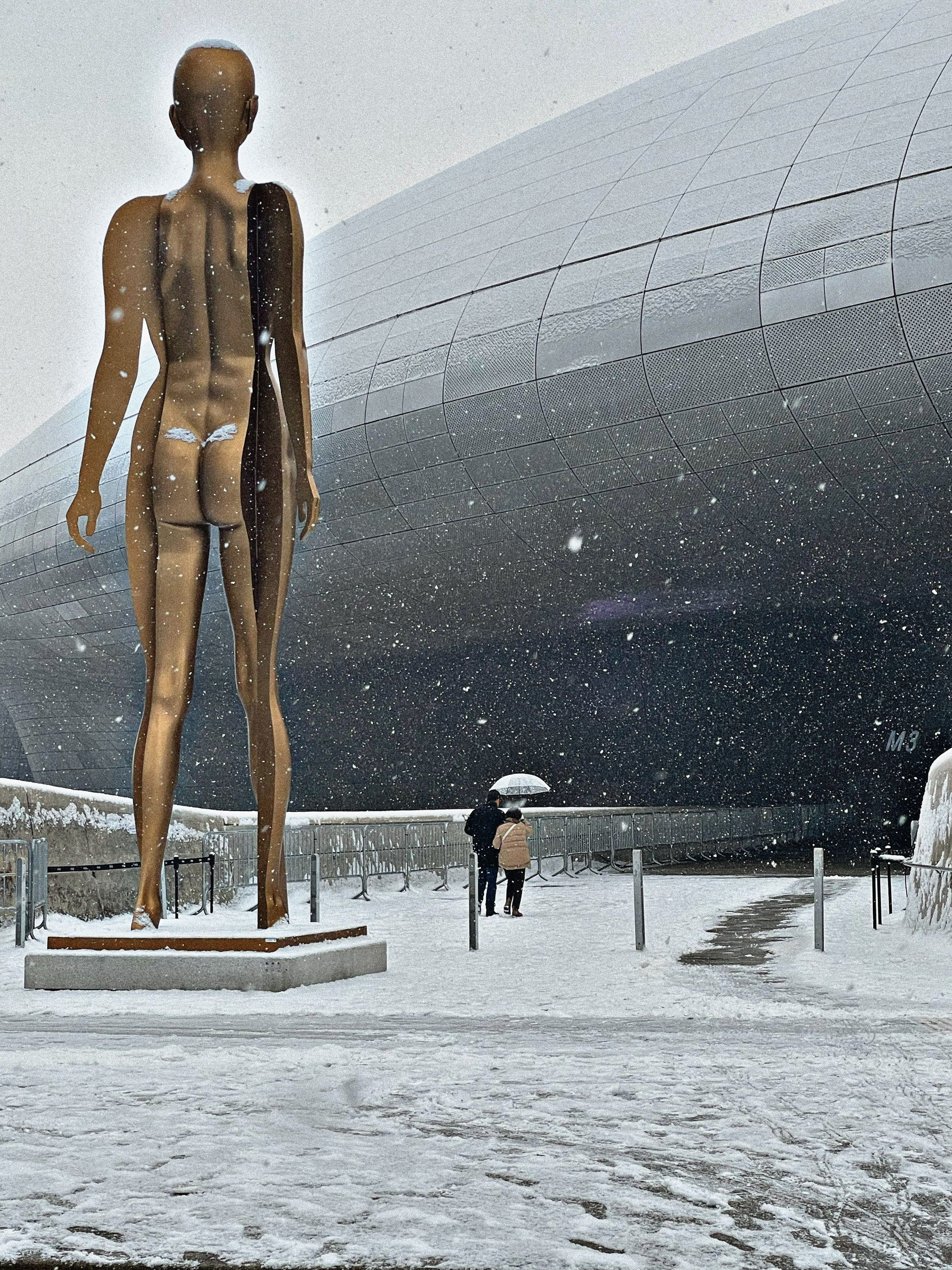 A tall sculpture of a naked lady beside the very futuristic dome of the Dongdaemun Design Plaza. Snow continues to fall.